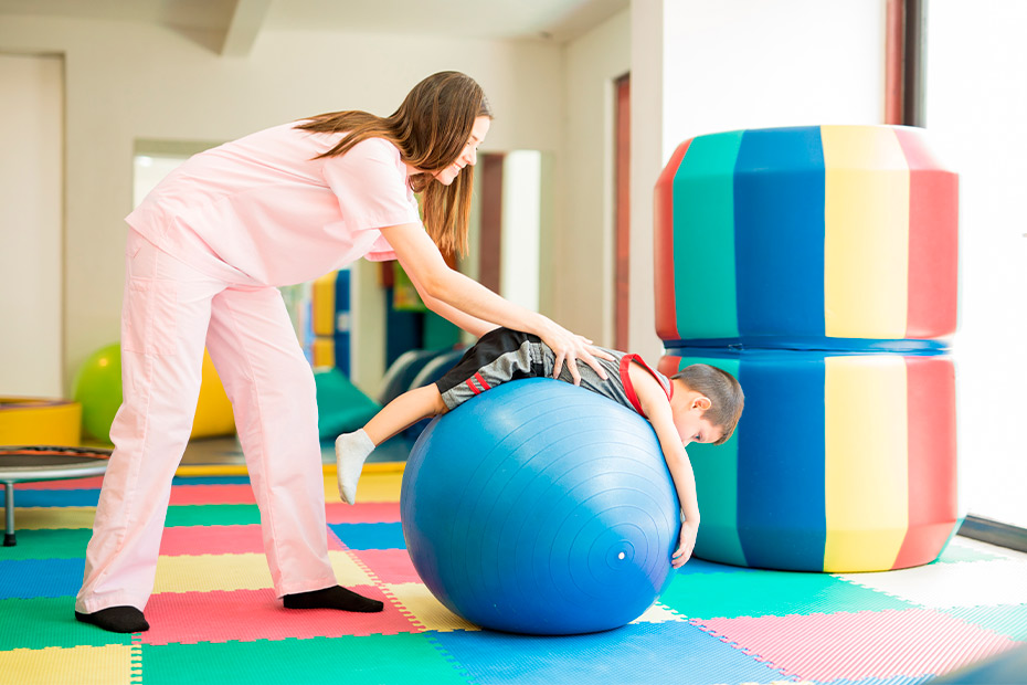 A woman supports a child on an exercise ball, illustrating the advantages of physical therapy for children with learning disabilities.