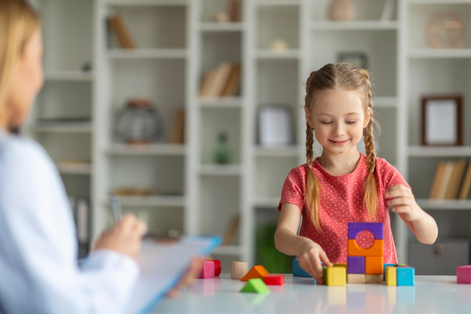 Cute little girl sitting at table and making pyramids from colorful wood blocks during meeting with psychotherapist, child development specialist lady looking at female kid and taking notes