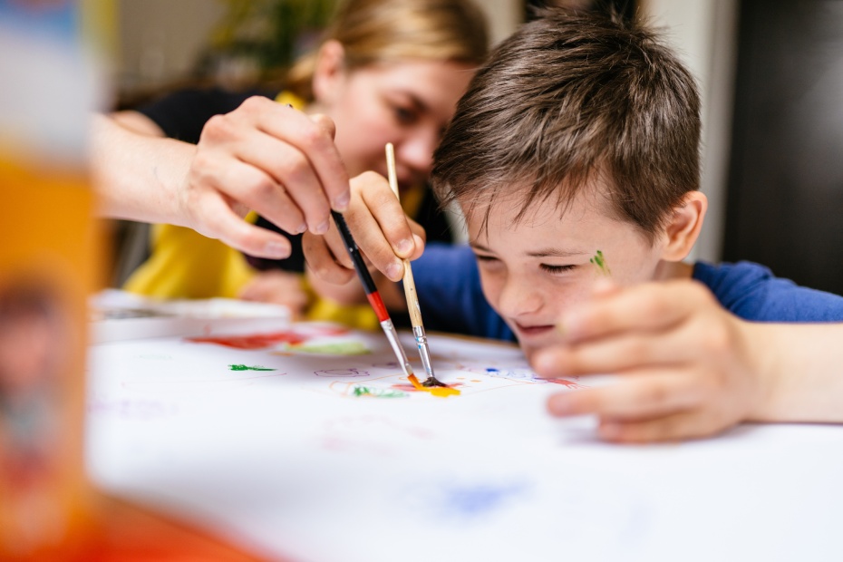 Young female teacher explain, talk at desk with a down syndrome schoolboy. Color painting for disabled kids, child who are down syndrome and student teacher, development of children with special needs