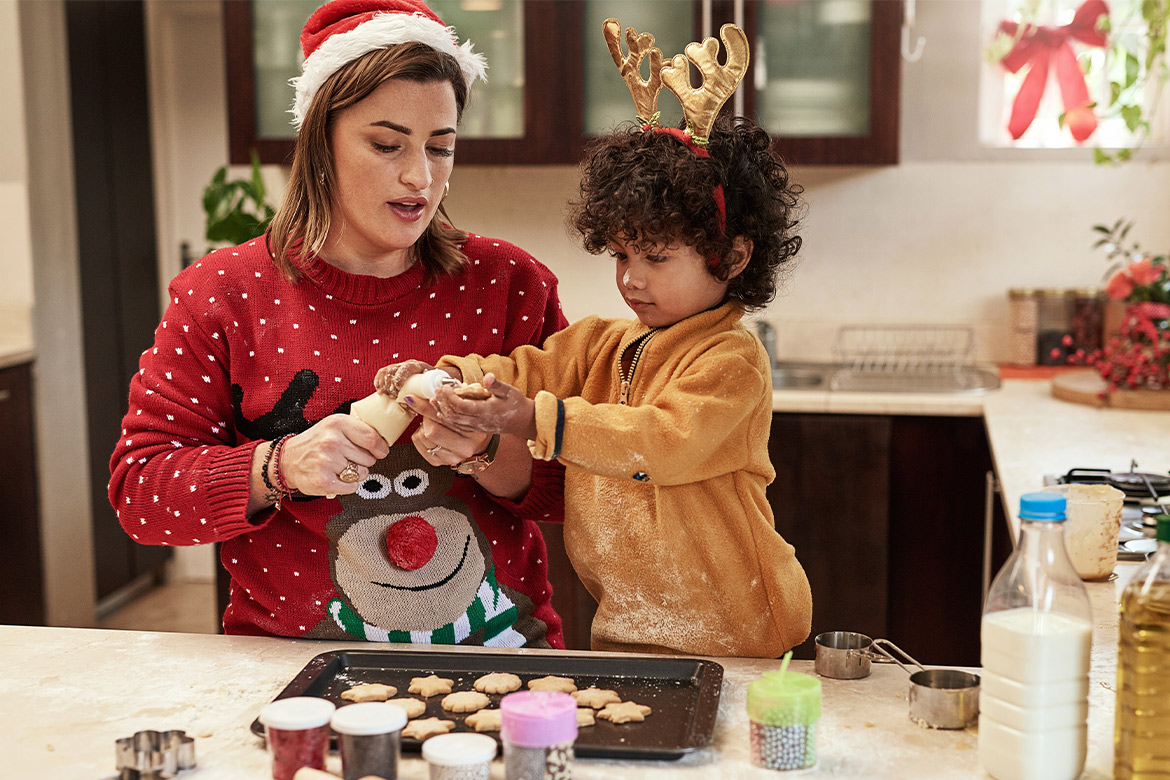 A woman and child dressed in Christmas outfits happily baking cookies, engaging in a fun activity to enhance motor skills.