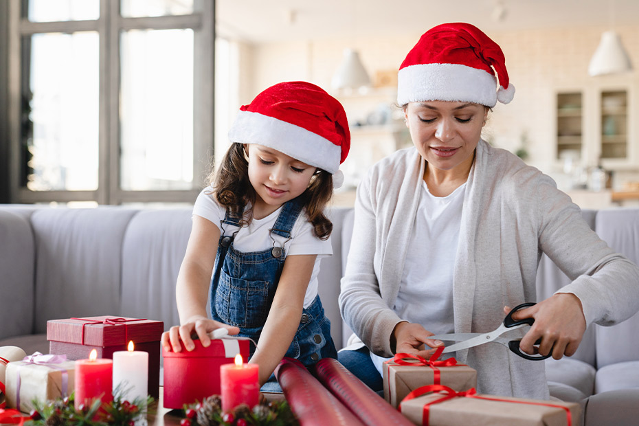 A woman and child wearing Santa hats are cutting gift wrap, engaging in a fun activity to enhance motor skills together.