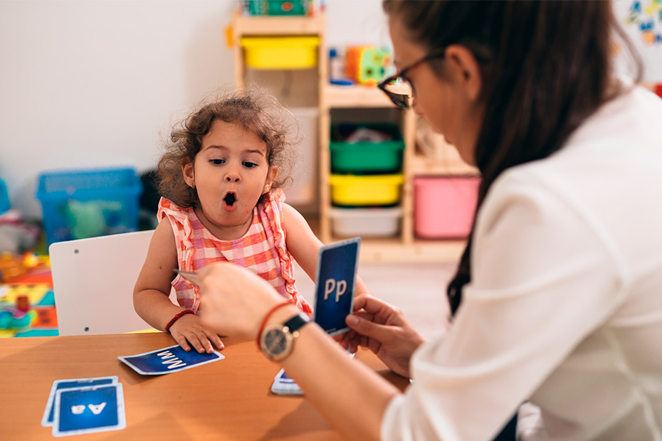 A woman engages playfully with a child in a classroom setting, highlighting interactive learning and speech therapy benefits.